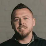 Professional headshot of smiling man with beard and short hair against gray background
