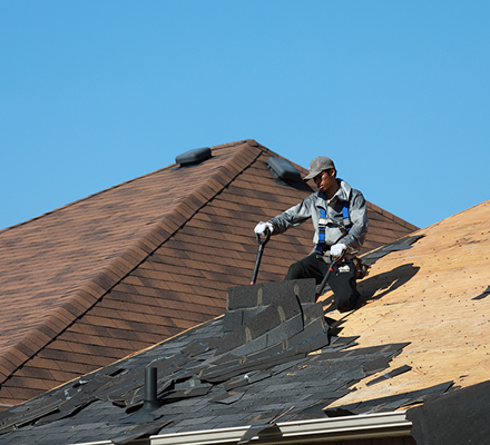 Roofer replacing shingles on a residential house roof under clear blue sky.
